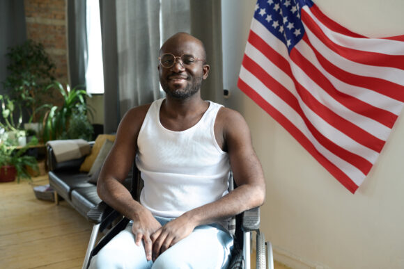 Portrait of African American man smiling at camera while sitting in wheelchair against the American flag on wall in the room