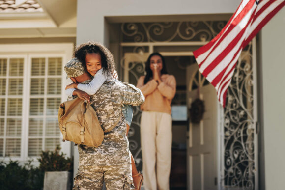 Servicemember returning home from service and hugging happy child. The servicemember's spouse stands in the background smiling.