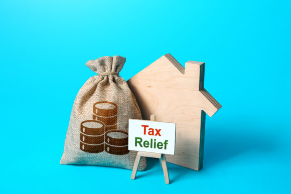 Wood cut out of a house next to a bag with coins on it with a sign that says "tax relief".